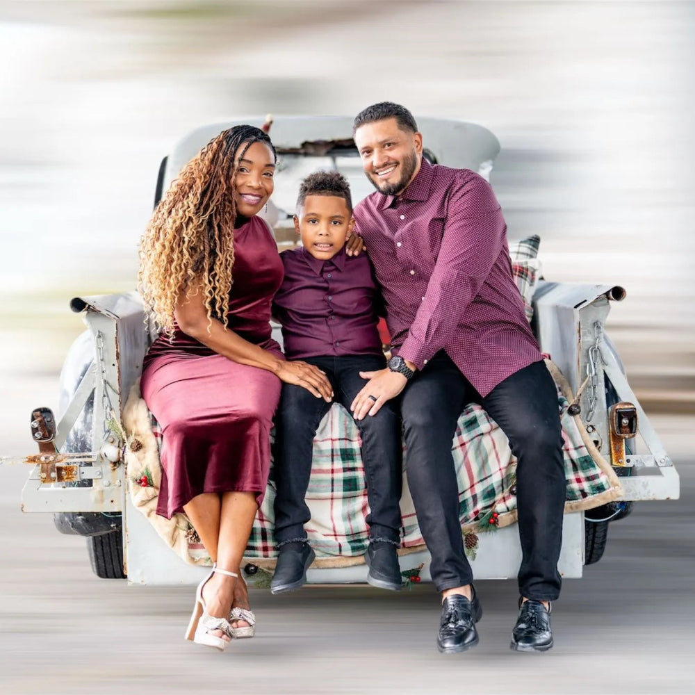 Family of three sitting on a vintage car with a plaid blanket, smiling at the camera.
